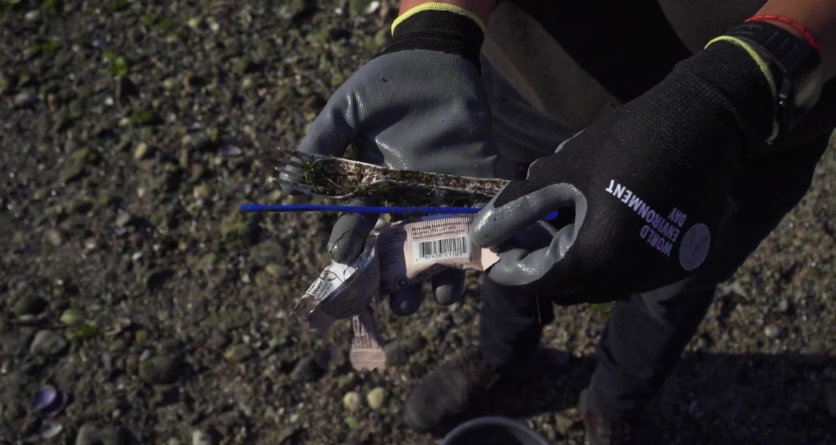 Deux mains portant des gants tiennent de petits déchets en plastique : une fourchette, une paille et un emballage de barre tendre. Sur un gant, il est écrit : "World Environment Day" (Journée mondiale de l'environnement)