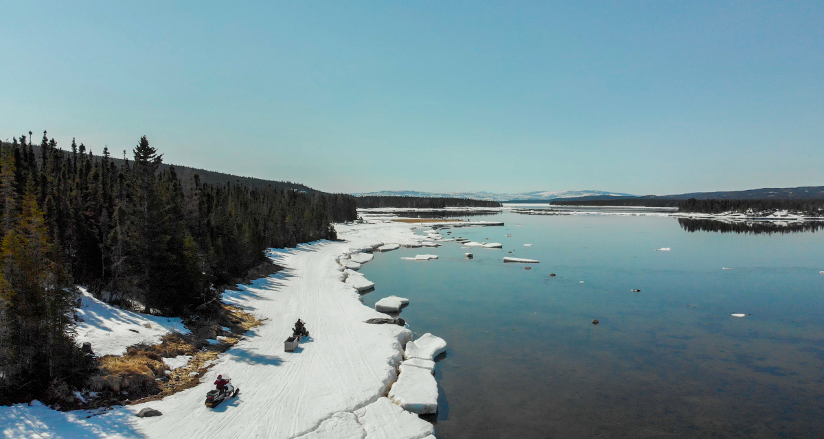 À Nunatsiavut, deux motoneigistes longent une forêt sur une partie gelée d'une rivière.