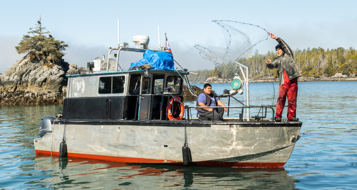 Two fishermen are on a boat in the middle of a lake. One is sitting and the other is casting a fishing net into the sea.