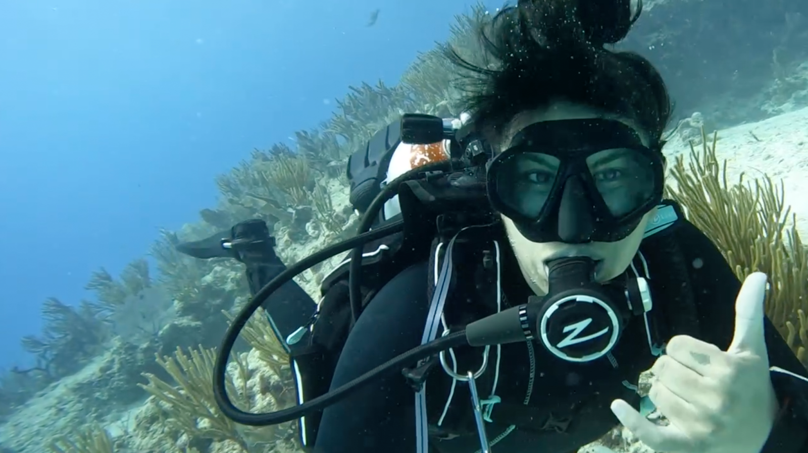 Alanna Canaran scuba diving wearing all black scuba gear. She is diving on a coral reef with different species of soft corals, like sea fans, and sea rods behind her.