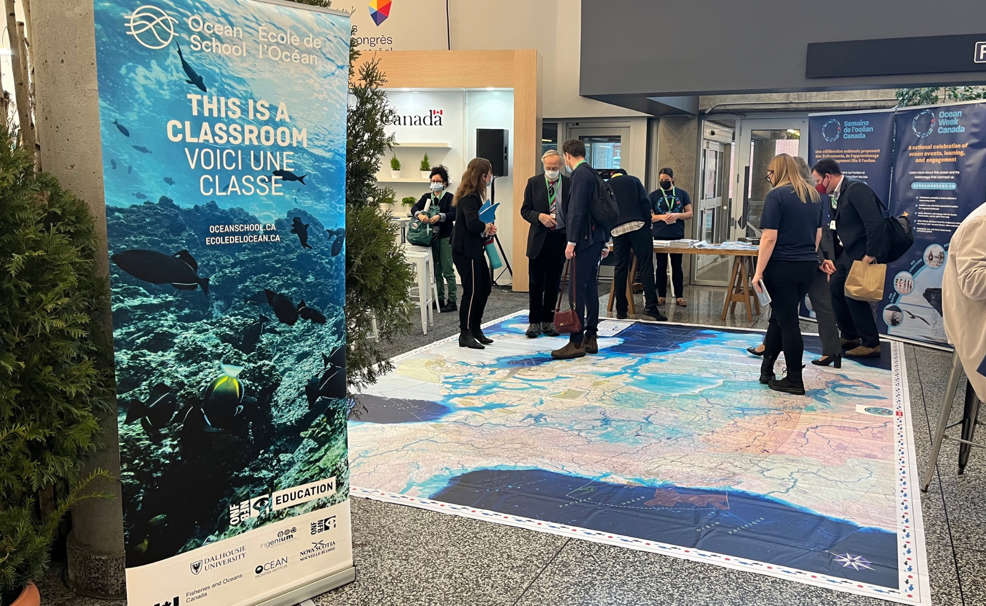 Photo of the Ocean, Freshwater and Us Giant Floor Map showing event at the Canadian Pavillion at COP 15. Attendees look down at the Giant-floor map and try it out. On the left side of the photo, there's an Ocean School standing banner. The banner features Ocean School logo and its partners' logo and the following text: "This is a classroom".