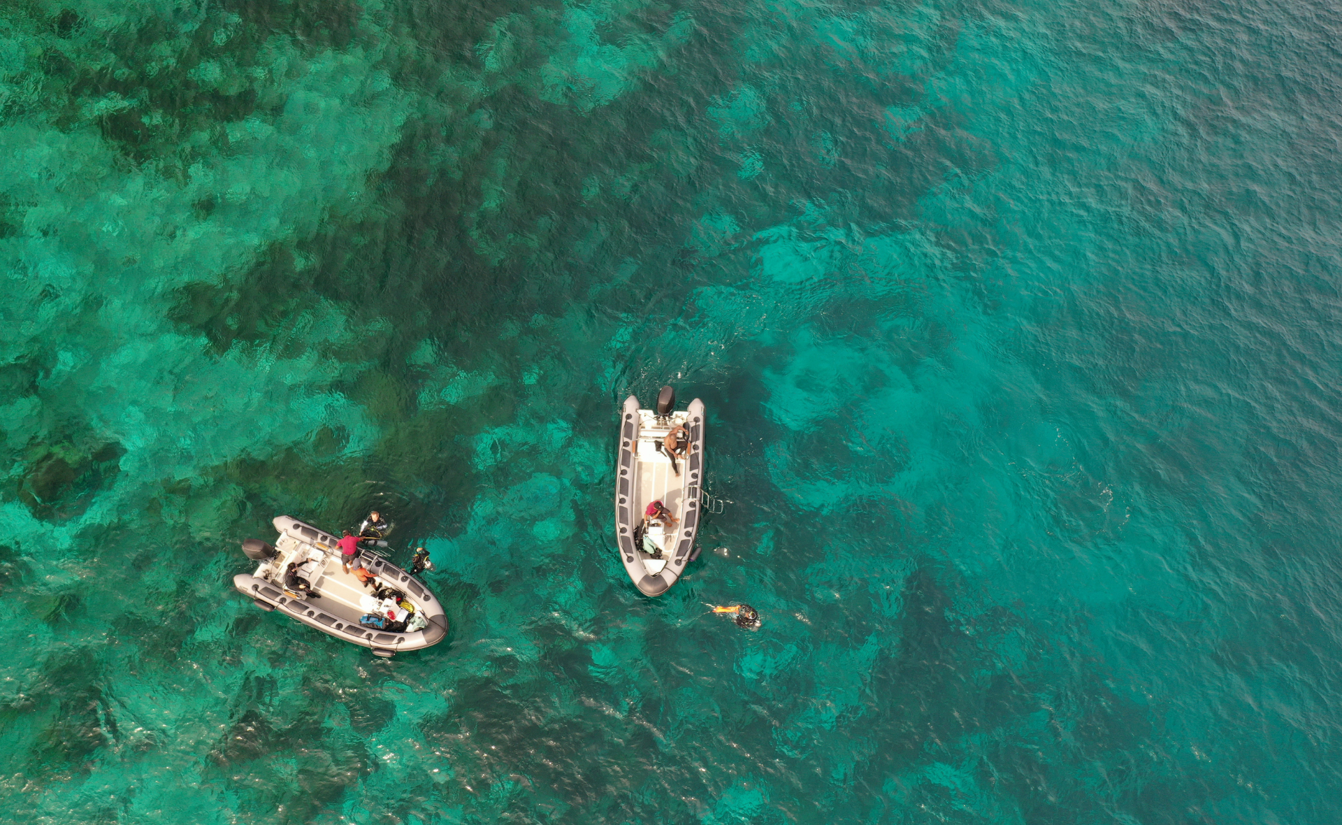 An image seen from above of two inflatable boats floating on clear turquoise water in Raja Ampat, Indonesia. The boats hold two and three people each and there are three divers swimming near them.