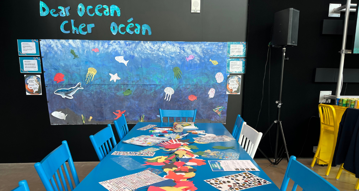 A blue table is covered with DIY materials. On the back wall, there's a painting on which drawings of sea creatures are pasted. Above the board is written: "Dear Ocean".