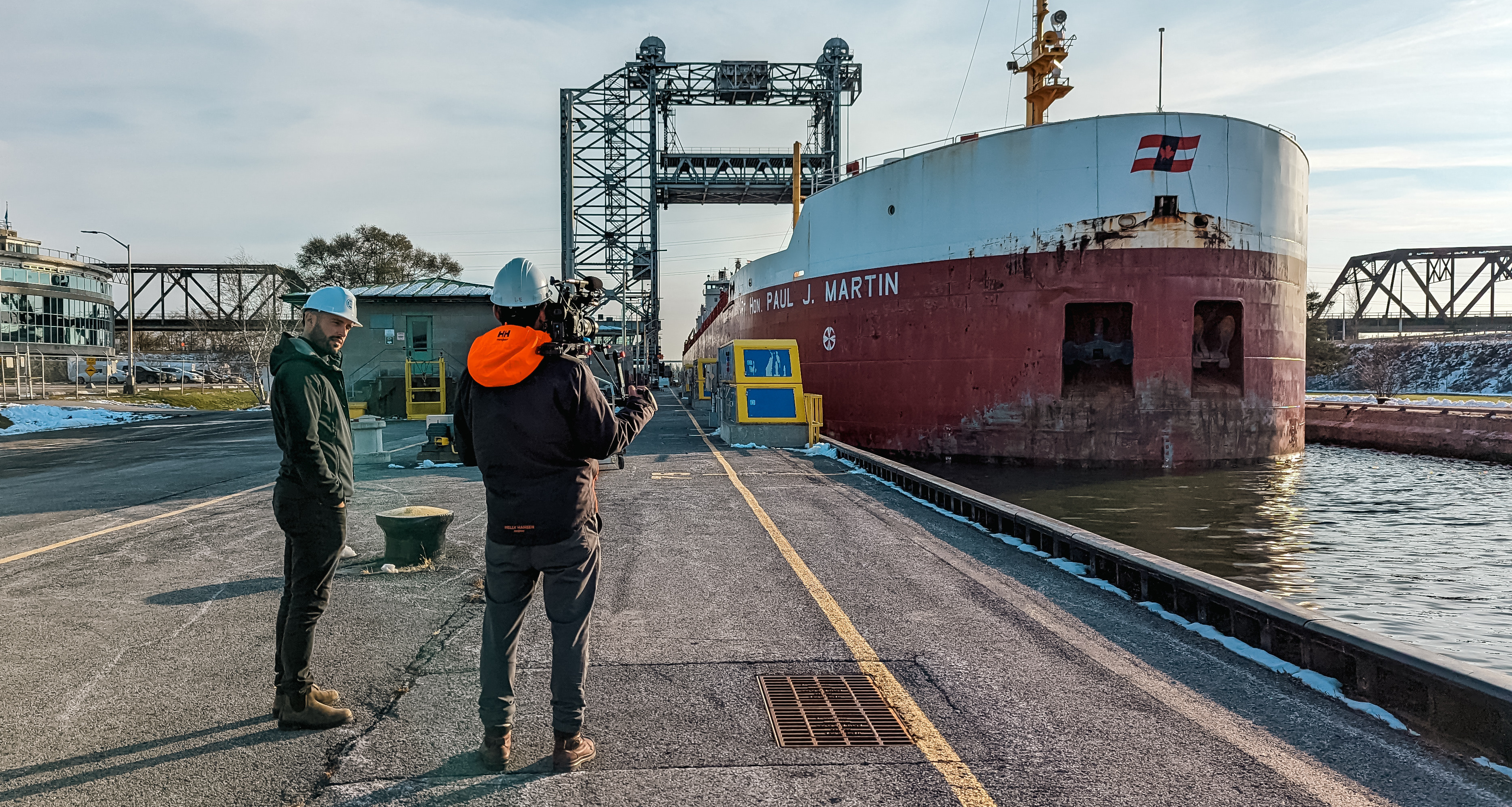 Le producteur Scott Simpson se tient sur la rive et pointe une caméra vers un grand cargo dans une écluse. Ricardo D'Amore se tient à côté de Scott.