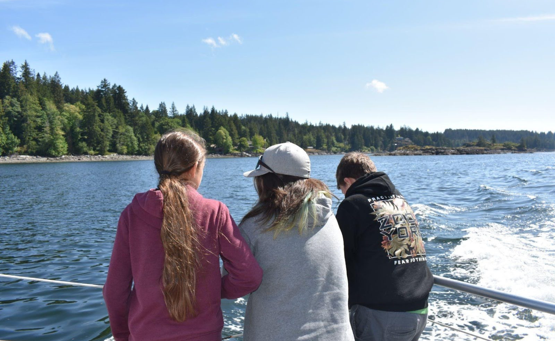 Three students stand on the deck of a boat looking over the water and trees in the distance.