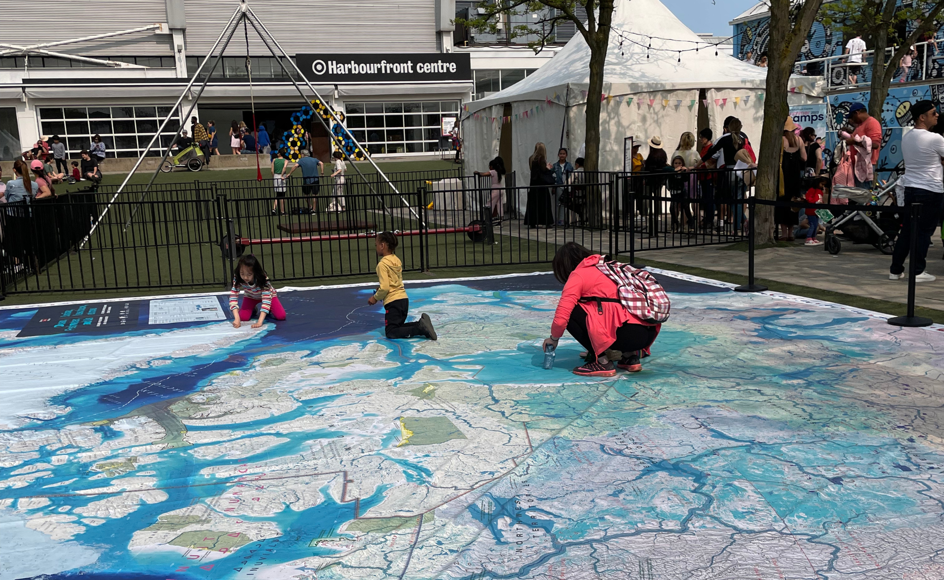 Un adulte et deux enfants sont accroupis et observent la carte-tapis géante du Canada. En arrière plan se trouve le Harbourfront centre de Toronto et d’autres kiosques et activités du JUNIOR festival.