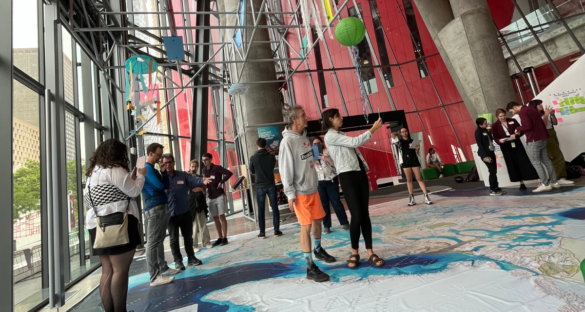 In the lobby of the National Film Board offices in Montreal, the Ocean School team shows university students how the giant-floor map The Ocean, Freshwater and Us works. Above the map, decorative jellyfish hang from scaffolding.