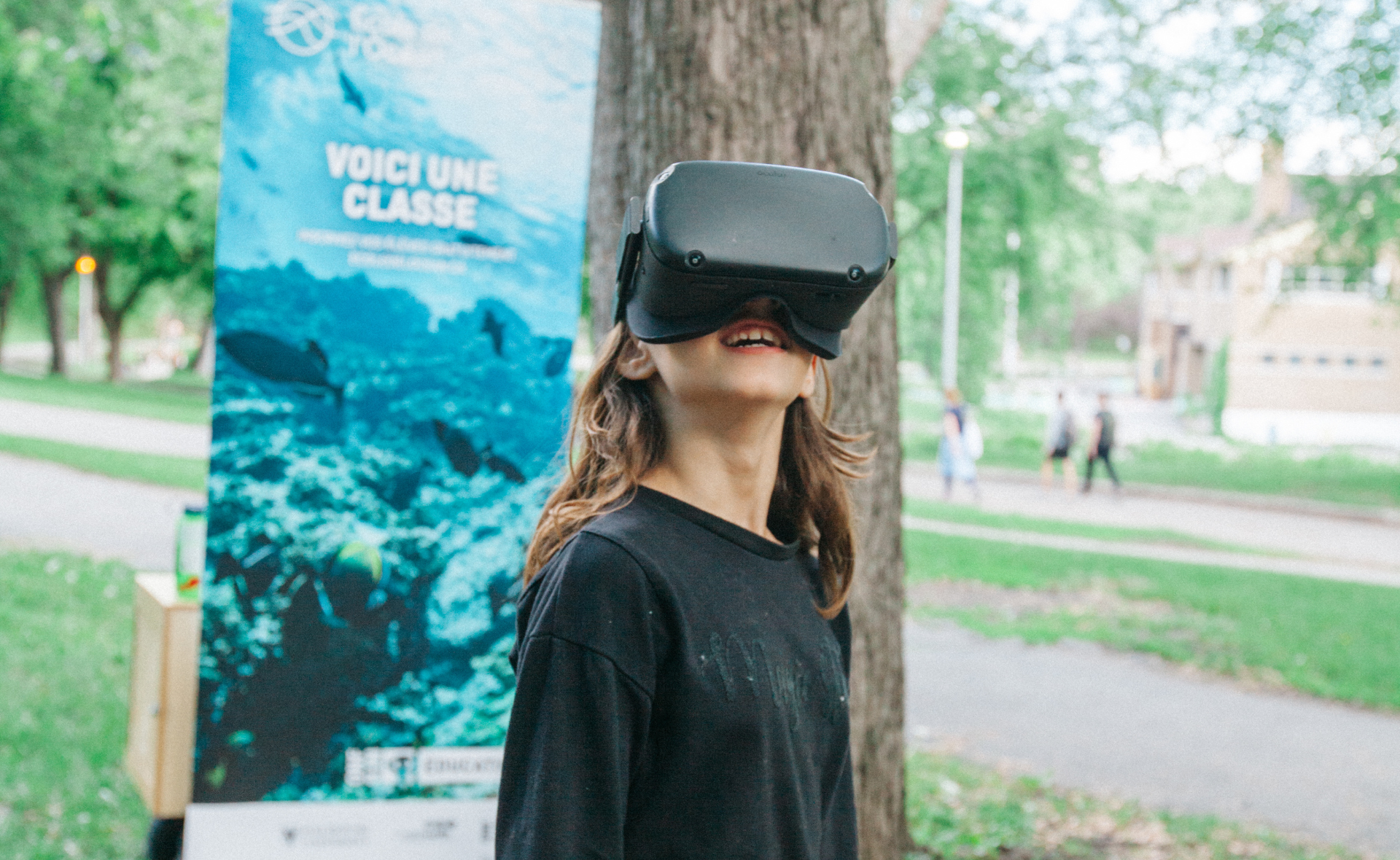 Outside, a child tries out Ocean School resources with a VR headset. In the background is an Ocean School promotional banner.
