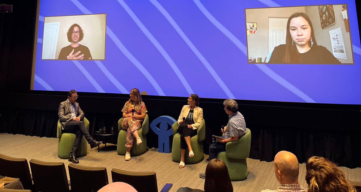 Three panellists and the panel moderator are seated on stage in a half-moon, facing the audience. Behind them, a screen projects two other panellists participating remotely.