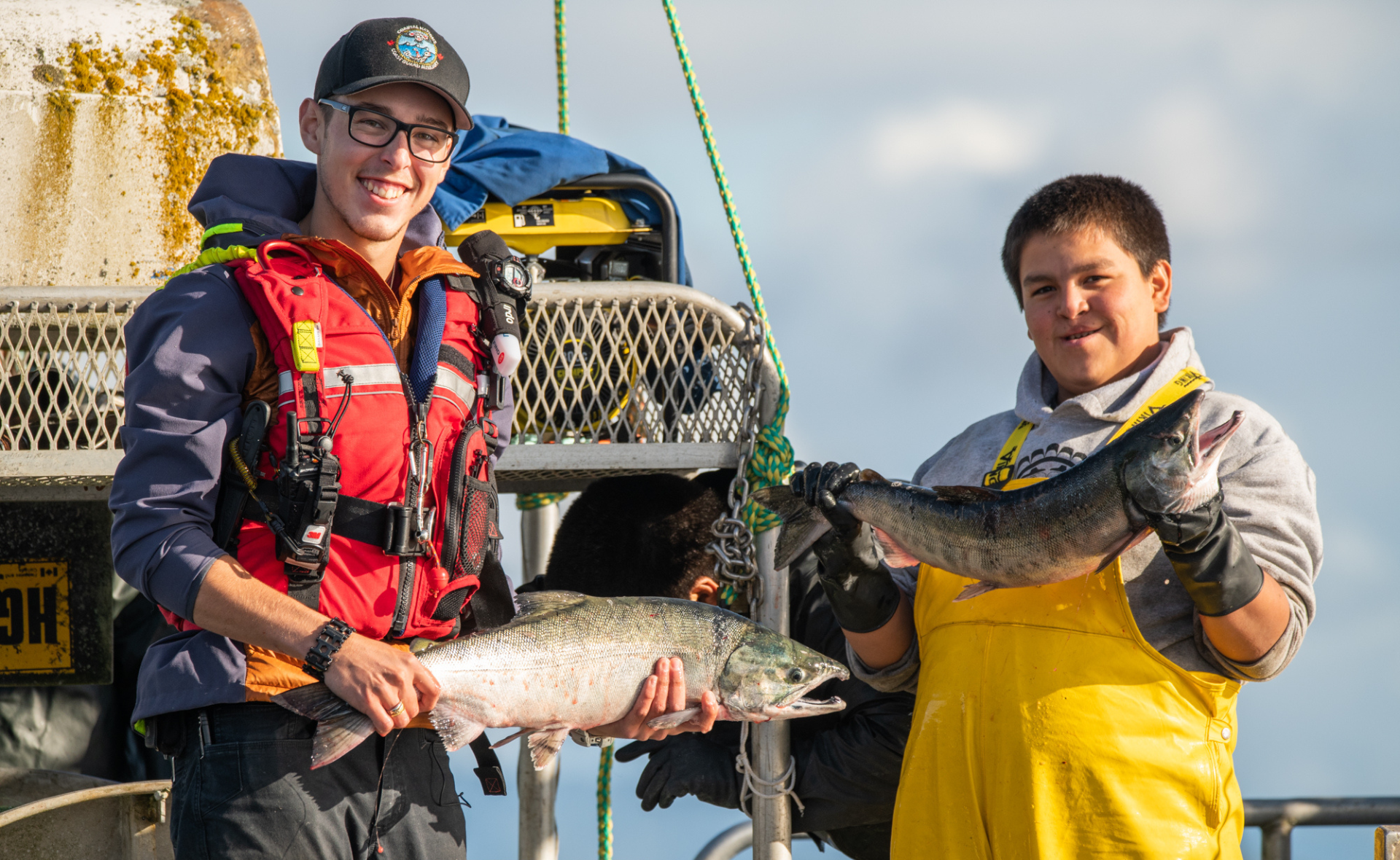 A promotional image for The Ocean School collection The Harvest. Two young Heiltsuk men hold up fish and smile for the camera.