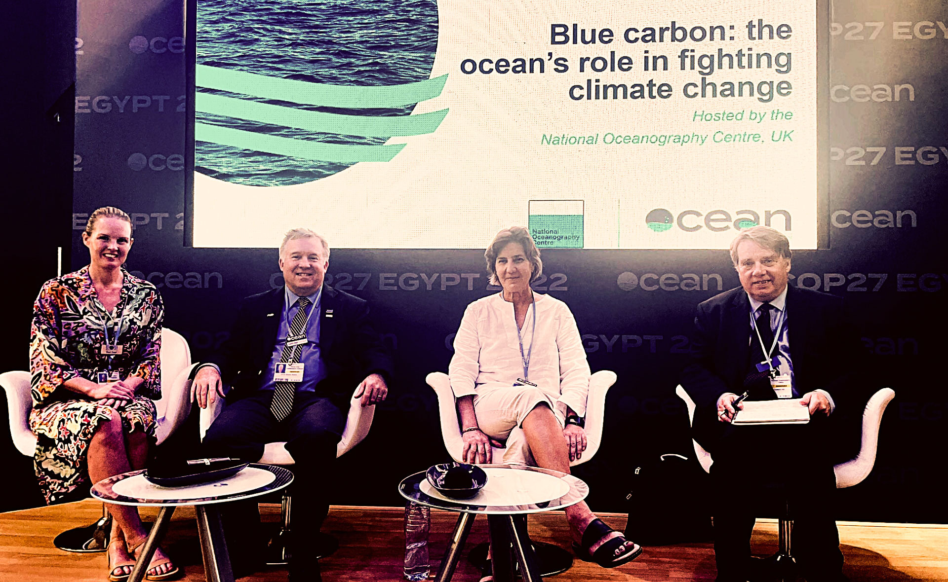 The four speakers of the panel discussion "Blue Carbon: The Ocean’s Role in Fighting Climate Change" at COP27 sitting in front of screen showing a slideshow. From left to right: Dr Narissa Bax (Researcher, University of Tasmania), Dr James Edson (Senior Scientist, WHOI), Dr Anya Waite (CEO of OFI Canada) and Professor Ed Hill, (Chief Executive Officer NOC).The following text is written on the slide behind them: Blue Carbon: The Ocean’s Role in Fighting Climate Change, hosted by the National Oceanography Centre, UK.