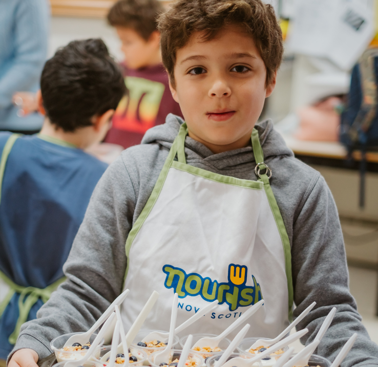 A child, wearing a Nourish apron, holds a tray full of yogurt dishes.