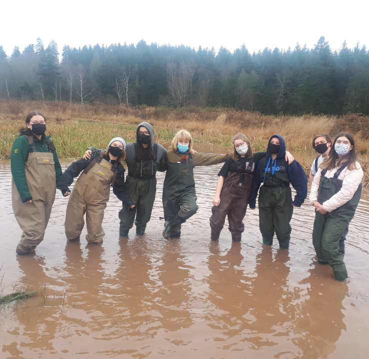 Michèle Banks stands arm-in-arm with her students in a pond. They are all wearing fisherman's overalls.