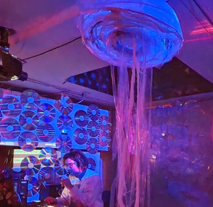 In a nightclub atmosphere, a DJ sits at his booth, next to a wall of vinyl records. In the foreground, a JellyTrash hangs from the ceiling.