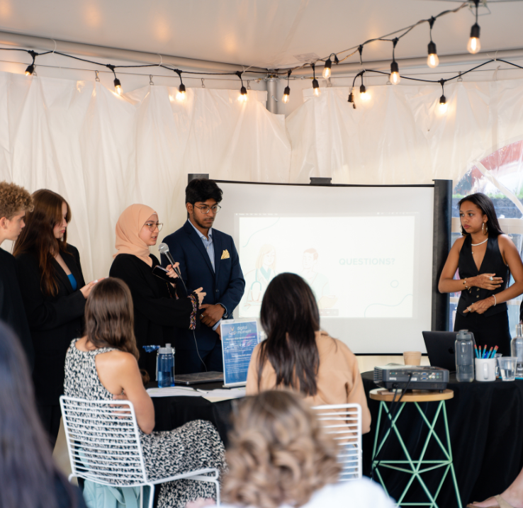 Under a big tent, five young people give a speech in front of a projection screen.