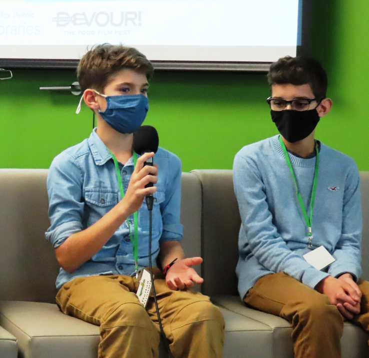 Two children are sitting on a couch wearing respirators. One of them is talking into a microphone.