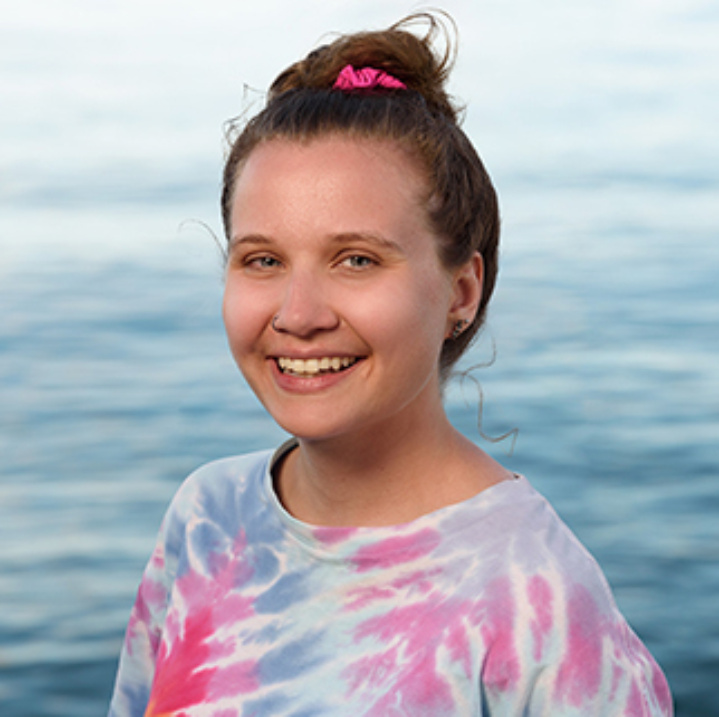A headshot of Alanna Akkermans. Alanna is smiling and wearing a blue and pink tie dye T-shirt. Her hair is tied into a bun with a pink scrunchie. There is a view of a body of water behind her.