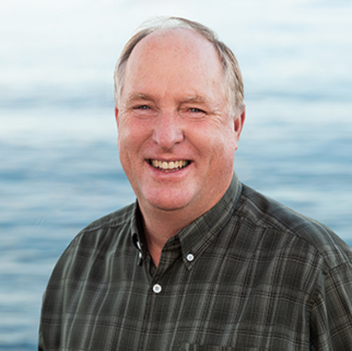 A headshot of Brian Hickey. Brian is smiling, wearing a green plaid dress shirt, and there is a view of a body of water behind him.