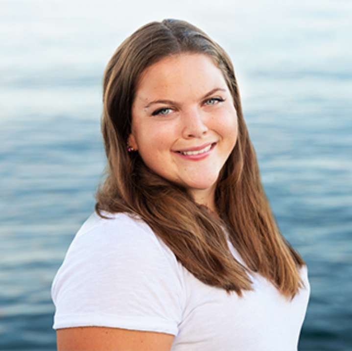 A headshot of Christina Charette. Christina is smiling and wearing a white T-shirt. There is a view of a body of water behind her.
