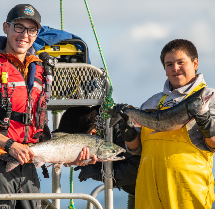 Une image promotionnelle pour la collection « La récolte » de l’École de l’Océan. Deux jeunes hommes de la nation Heiltsuk tiennent des poissons et sourient à la caméra.