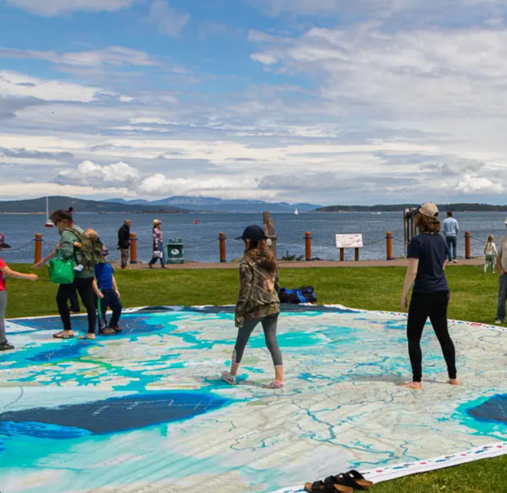 Near a body of water, outside, people are trying out the giant floor map