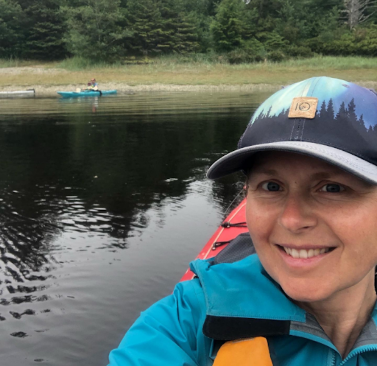 Kim smiles at the camera while sitting in a kayak on a body of water with a shoreline in the distance.